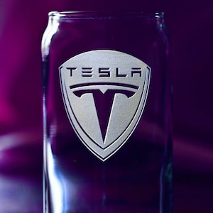 May include: A clear glass drinking glass with the Tesla logo etched on the front. The logo is silver and features the Tesla 'T' in a shield shape.