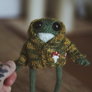 Peut inclure: Une grenouille en tricot vert portant un pull à capuche jaune et vert avec un motif de champignon. La grenouille a de grands yeux noirs et une bouche blanche.