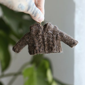 Puede incluir: Un pequeño cárdigan de punto marrón con un patrón de punto de cable y botones dorados.