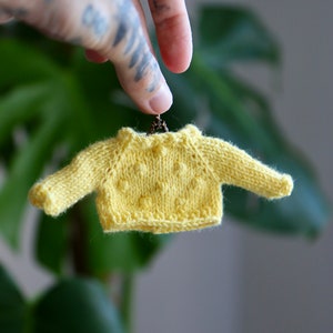 May include: A small yellow knitted sweater with a textured pattern hangs from a chain. The sweater is about the size of a doll's outfit.