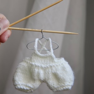 Peut inclure: Un body bébé blanc en tricot avec un design de bretelles croisées, suspendu à un cintre en fil de fer. Le body est fait d'un fil doux et texturé et a une bordure de dentelle délicate.