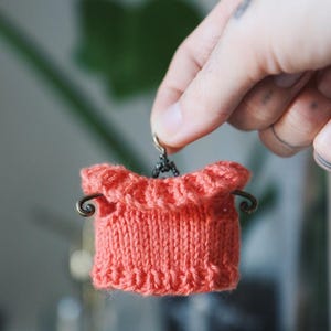 May include: A small, coral-colored knitted item with a ruffled top edge and decorative metal accents. The item is held by a hand, showcasing its intricate knit texture and design. The piece appears to be a miniature garment or accessory.