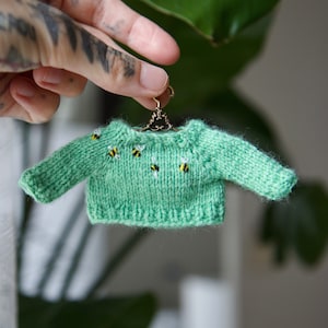 Peut inclure: Un mini pull en tricot vert avec un petit cintre et des abeilles brodées.