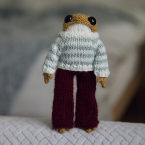 Könnte beinhalten: Eine gestrickte Froschpuppe, die einen gestreiften Pullover und eine braune Hose trägt. Die Puppe steht auf einer weißen, strukturierten Oberfläche.