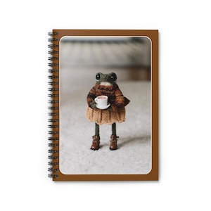 Peut inclure: Cahier à spirales avec une image fantaisiste d'une poupée grenouille. La grenouille porte un pull, une jupe et des bottes marron et beige, et tient une petite tasse blanche. Le cahier a un bord marron et une reliure à spirales.