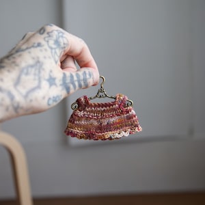 May include: A small, hand-knitted dress in shades of red, brown, and cream, hanging on a miniature gold hanger.