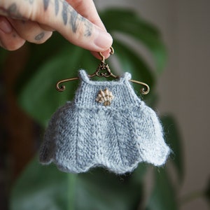 Peut inclure: Une petite robe en tricot gris avec un ourlet festonné, suspendue à un petit cintre de style antique avec un motif de volutes décoratif. La robe a un petit bouton rond de couleur or sur le devant.