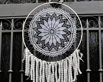 Puede incluir: Un atrapasueños de crochet blanco con pompones blancos y encaje blanco que cuelgan de un aro dorado. El atrapasueños está colgado de una puerta de metal negro.