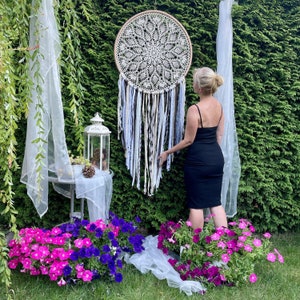Puede incluir: Un gran atrapasueños de crochet blanco con plumas marrones y blancas que cuelgan de él. El atrapasueños está colgado frente a un arbusto verde y una mujer está de pie detrás de él.