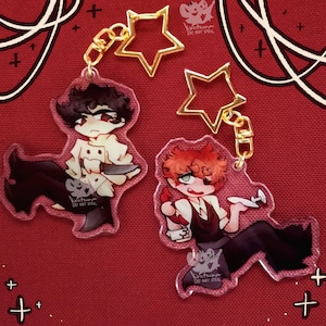 Op de afbeelding: Twee karaktercharms aan een gouden ketting met stervormige sleutelhangers. De charms zijn van helder acryl met illustraties van twee personages, een met zwart haar en een wit shirt en de ander met rood haar en een zwart shirt. De tekst "Kratsun DO NOT STEAL" staat op elke charm gedrukt.
