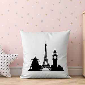 May include: A white square pillow with a black silhouette of famous landmarks, including the Eiffel Tower, a pagoda, and Big Ben. The pillow is on a wooden surface, with a star-shaped pillow and a shelf in the background.