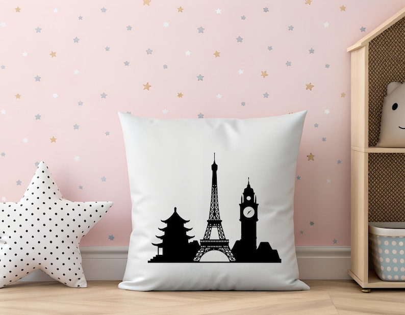 May include: A white square pillow with a black silhouette of famous landmarks, including the Eiffel Tower, a pagoda, and Big Ben. The pillow is on a wooden surface, with a star-shaped pillow and a shelf in the background.