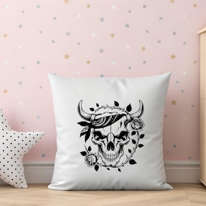 May include: White square pillow featuring a black and white skull design. The skull has horns, a bandana, and is surrounded by roses and thorns. The pillow is on a wooden surface in front of a pink wall with star-shaped decorations.