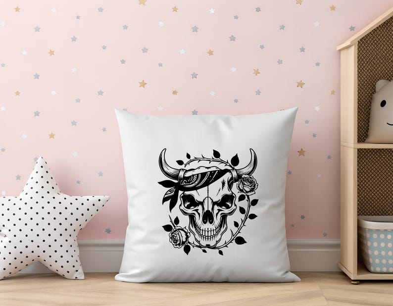 May include: White square pillow featuring a black and white skull design. The skull has horns, a bandana, and is surrounded by roses and thorns. The pillow is on a wooden surface in front of a pink wall with star-shaped decorations.
