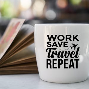 May include: A white ceramic mug with the words "WORK SAVE TRAVEL REPEAT" in black. A small airplane graphic is included. An open book with colorful pages is in the background. The mug is on a white surface.