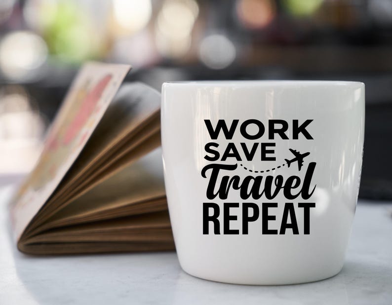 May include: A white ceramic mug with the words "WORK SAVE TRAVEL REPEAT" in black. A small airplane graphic is included. An open book with colorful pages is in the background. The mug is on a white surface.