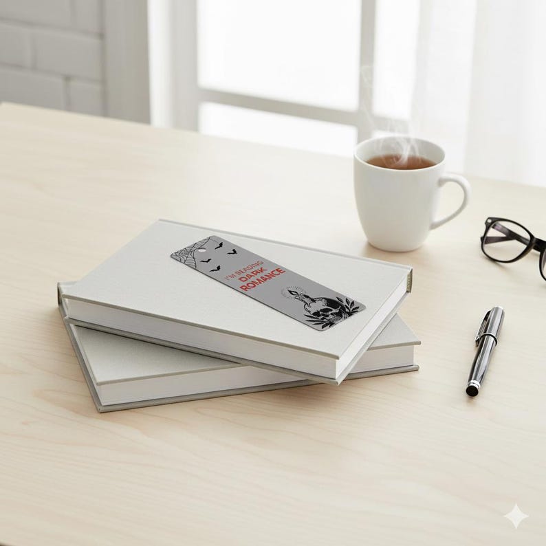 May include: Two white books are stacked on a light wood surface, with a gray bookmark featuring a skull and the text "I'M READING DARK ROMANCE." A white mug with tea, glasses, and a pen are also on the table.