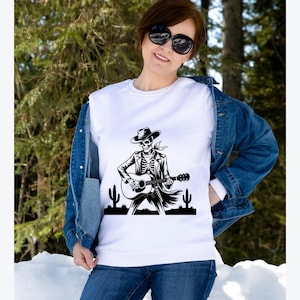 May include: White sweatshirt featuring a black and white graphic of a skeleton playing a guitar, wearing a cowboy hat and jacket. The design includes cacti silhouettes. The sweatshirt is worn with a denim jacket and blue jeans.