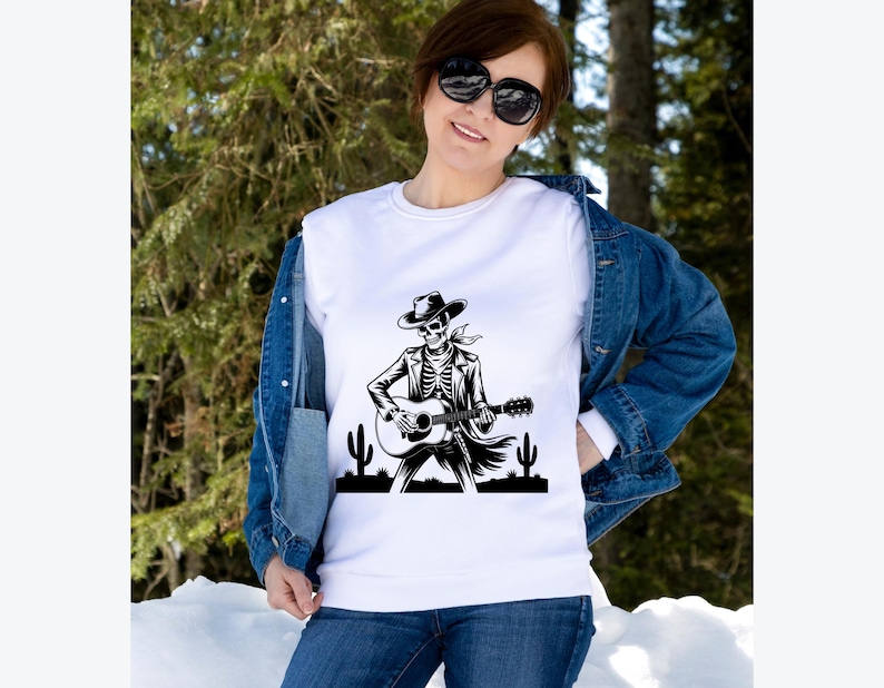 May include: White sweatshirt featuring a black and white graphic of a skeleton playing a guitar, wearing a cowboy hat and jacket. The design includes cacti silhouettes. The sweatshirt is worn with a denim jacket and blue jeans.