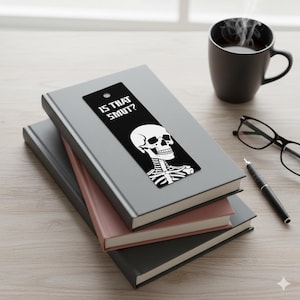 May include: A black bookmark with a white skull graphic and the text "IS THAT SMUT?" rests on a stack of books. A black coffee mug, glasses, and a pen are also on the wooden surface.