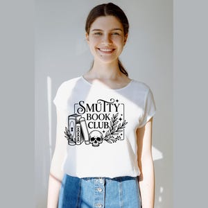 May include: White t-shirt with a black graphic that reads "Smutty Book Club." The design includes two books, a skull, and botanical elements. The shirt is made of a soft material and has a crew neck.