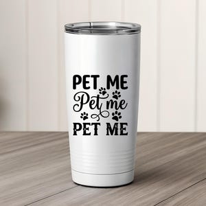 May include: A white tumbler with a silver rim and a black "PET ME" design, including paw prints. The text is in a mix of fonts. The tumbler is cylindrical and sits on a wooden surface.