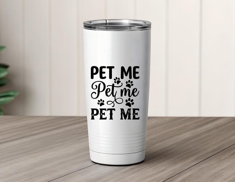 May include: A white tumbler with a silver rim and a black "PET ME" design, including paw prints. The text is in a mix of fonts. The tumbler is cylindrical and sits on a wooden surface.