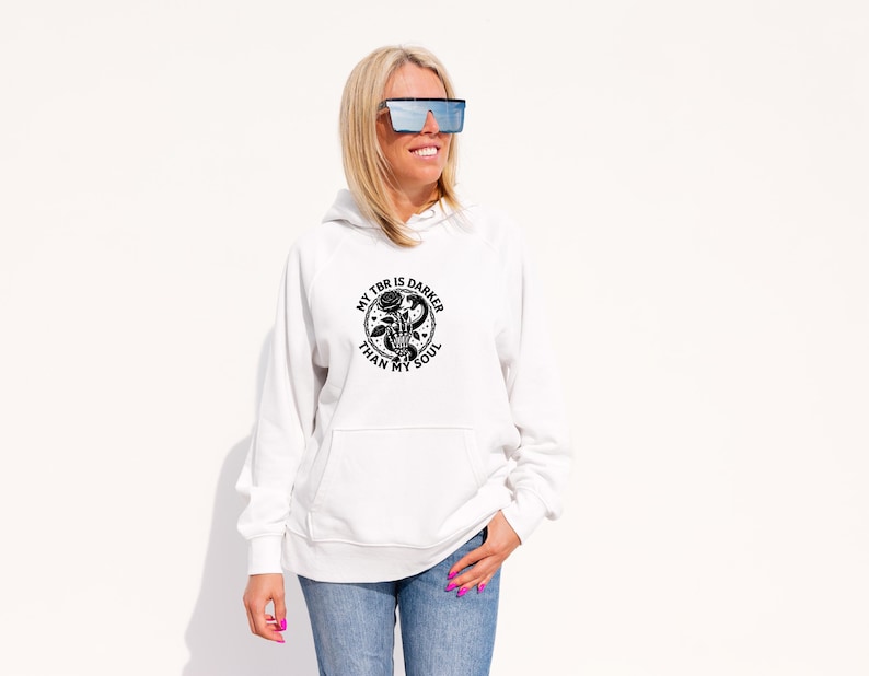 May include: White hoodie with a front pocket and a black graphic that reads "MY TBR IS DARKER THAN MY SOUL" with a rose and hand design. The person is wearing blue jeans and sunglasses.