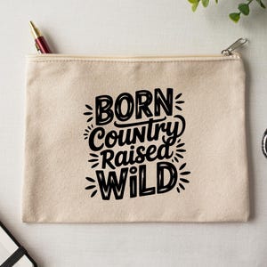 May include: A beige canvas zipper pouch with the text "BORN Country RAISED WILD" in black. A red and gold pen is in the zipper. The pouch is on a white surface with scissors and greenery.