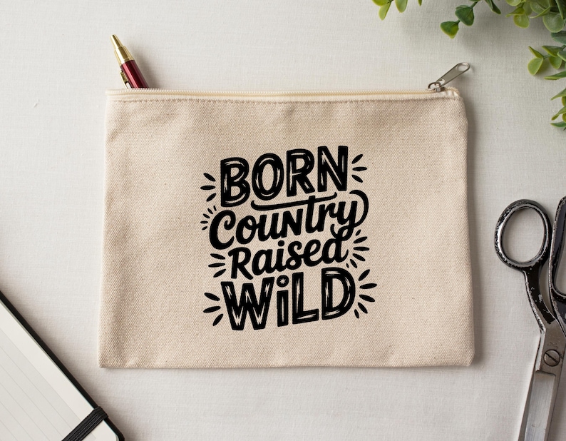 May include: A beige canvas zipper pouch with the text "BORN Country RAISED WILD" in black. A red and gold pen is in the zipper. The pouch is on a white surface with scissors and greenery.