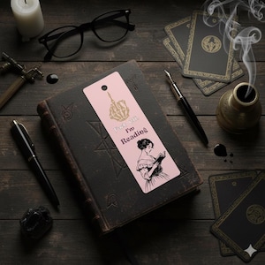 May include: A pink bookmark with the text "Fuck Off, I'm Reading" and a skeleton hand illustration. The bookmark also features a black and white illustration of a woman reading. The bookmark is on top of a dark book.