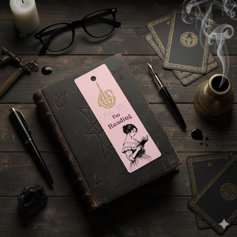 May include: A pink bookmark with the text "Fuck Off, I'm Reading" and a skeleton hand illustration. The bookmark also features a black and white illustration of a woman reading. The bookmark is on top of a dark book.