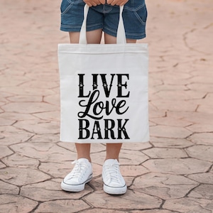 May include: A white tote bag with the words "LIVE Love BARK" in black script. The bag is held by a person wearing denim shorts and white sneakers. The background is a textured, brown surface.