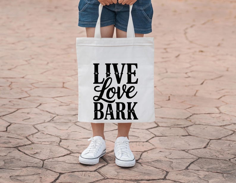 May include: A white tote bag with the words "LIVE Love BARK" in black script. The bag is held by a person wearing denim shorts and white sneakers. The background is a textured, brown surface.