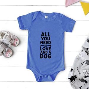 May include: A blue baby onesie with the text "ALL YOU NEED IS LOVE AND A DOG" in black. The onesie is next to a pair of gray baby slippers with pink accents and a patterned blanket.