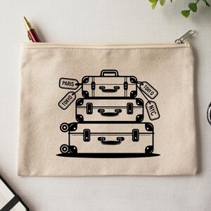 May include: A beige canvas zipper pouch featuring a black illustration of stacked vintage suitcases with tags that read "Paris", "Tokyo", and "NYC". The design includes handles, latches, and wheels on the bottom suitcase.