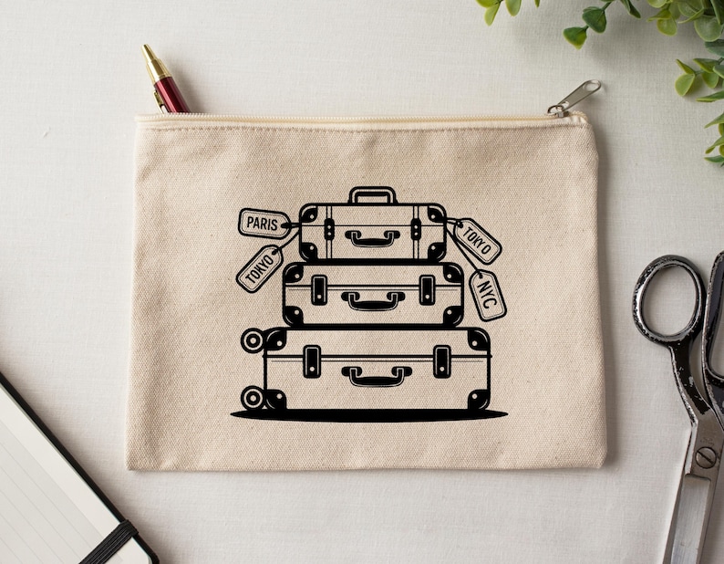 May include: A beige canvas zipper pouch featuring a black illustration of stacked vintage suitcases with tags that read "Paris", "Tokyo", and "NYC". The design includes handles, latches, and wheels on the bottom suitcase.