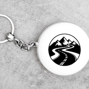May include: A white round keychain with a black and white mountain road design. The keychain has a silver metal ring and chain. The design features a winding road leading towards mountain peaks.