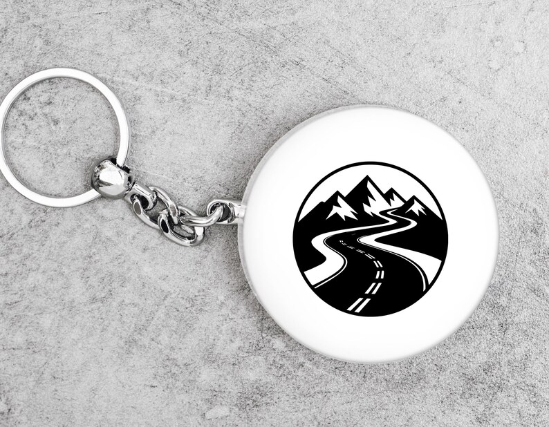 May include: A white round keychain with a black and white mountain road design. The keychain has a silver metal ring and chain. The design features a winding road leading towards mountain peaks.