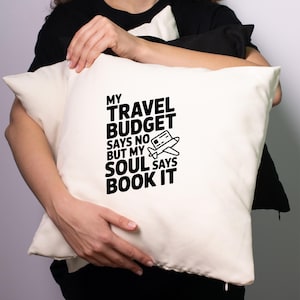 May include: Cream-colored pillow with the text "MY TRAVEL BUDGET SAYS NO BUT MY SOUL SAYS BOOK IT" in black. A small airplane graphic is included. The pillow is rectangular and appears to be made of a soft fabric.