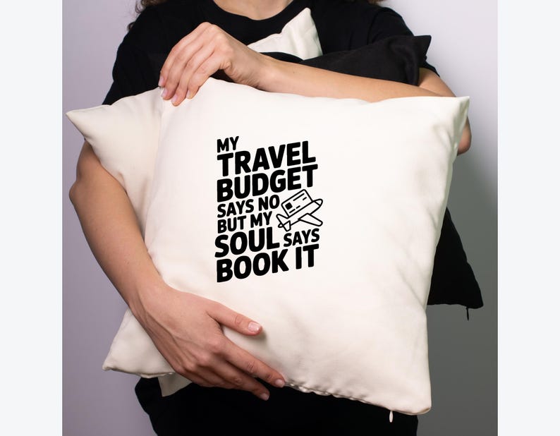 May include: Cream-colored pillow with the text "MY TRAVEL BUDGET SAYS NO BUT MY SOUL SAYS BOOK IT" in black. A small airplane graphic is included. The pillow is rectangular and appears to be made of a soft fabric.