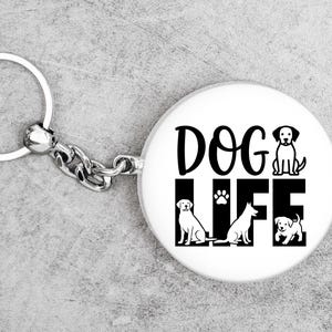 May include: White keychain with a round pendant featuring the words "DOG LIFE" in black, along with illustrations of three dogs and a paw print. The keychain has a silver ring and chain.