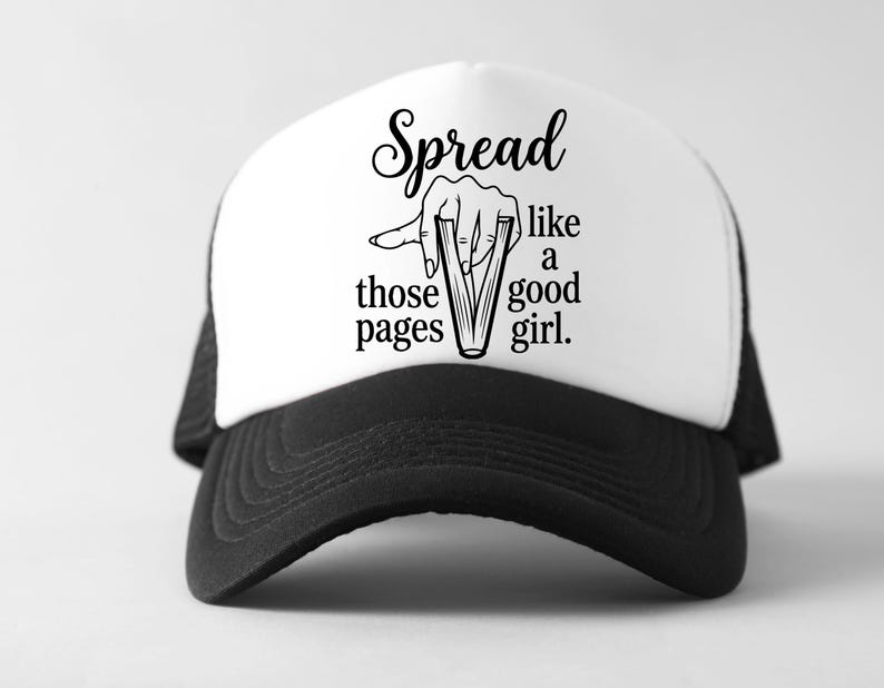 May include: A black and white trucker hat with the text "Spread like those pages like a good girl." The hat has a white front panel and a black brim and mesh back. The graphic shows a hand holding open pages.