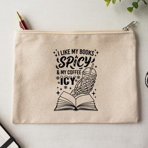 May include: A beige canvas zipper pouch with the text "I LIKE MY BOOKS SPICY & MY COFFEE ICY" in black. A skeleton hand holds an iced coffee over an open book. A pen is visible in the zipper.
