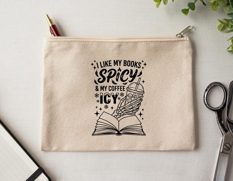 May include: A beige canvas zipper pouch with the text "I LIKE MY BOOKS SPICY & MY COFFEE ICY" in black. A skeleton hand holds an iced coffee over an open book. A pen is visible in the zipper.