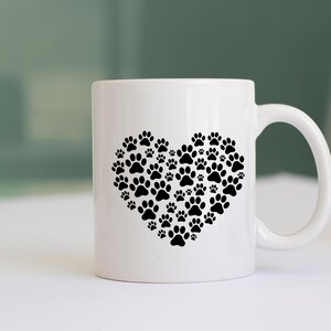 May include: White ceramic mug featuring a heart design made of black paw prints. The mug has a curved handle and is suitable for hot or cold beverages. A perfect gift for pet lovers.