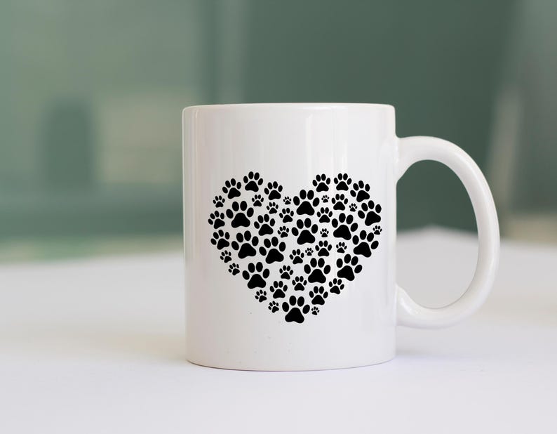 May include: White ceramic mug featuring a heart design made of black paw prints. The mug has a curved handle and is suitable for hot or cold beverages. A perfect gift for pet lovers.