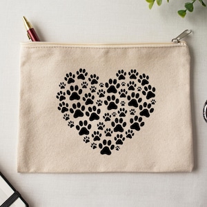 May include: A beige canvas zippered pouch featuring a heart made of black paw prints. A red and gold pen is visible in the zipper opening. The pouch is on a white surface.