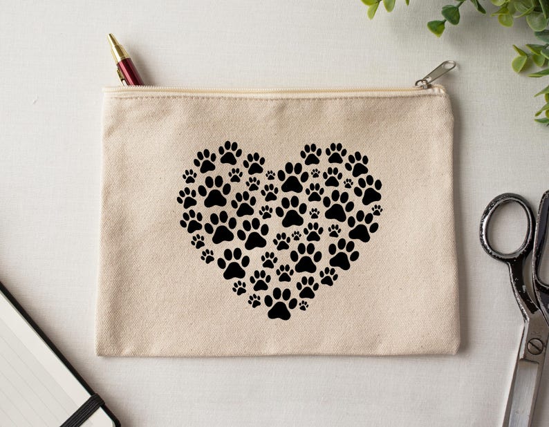 May include: A beige canvas zippered pouch featuring a heart made of black paw prints. A red and gold pen is visible in the zipper opening. The pouch is on a white surface.