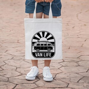 May include: A white tote bag featuring a black and white graphic of a vintage van with surfboards, the sun, and the words "VAN LIFE". The bag is held by a person wearing denim shorts and white sneakers.